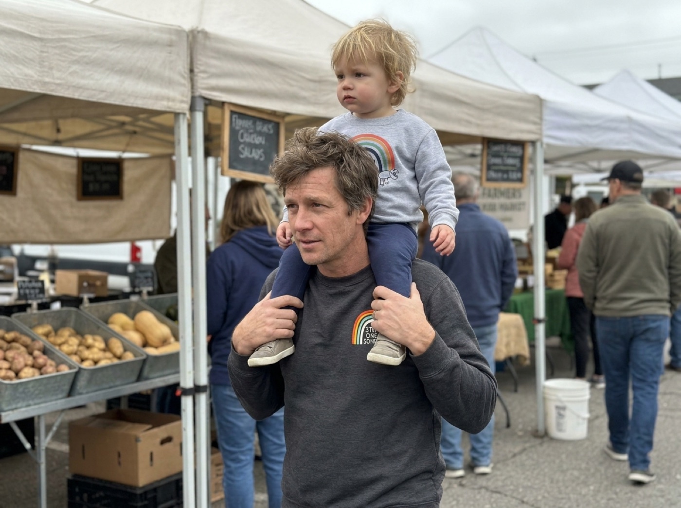 A dad in a rainbow crewneck carries his toddler in a rainbow sweatshirt on his shoulders at a farmers market